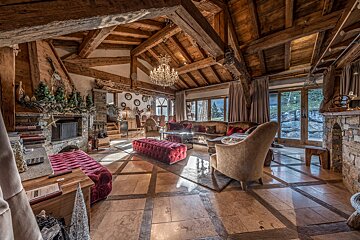 Grand chalet living room with exposed wooden beams, stone fireplaces, plush red velvet seating, a chandelier, and sunny tiled floors.