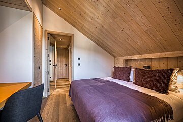 Cozy bedroom with a sloped wooden ceiling, light wood accents, and a bed with a purple blanket. A desk and chair are visible on the left.