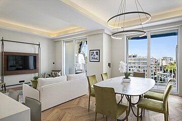 Modern open-plan apartment with a white sofa, olive green dining chairs, chic chandelier, and sliding glass doors to a balcony.