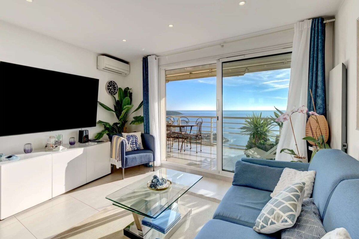 Sunny blue living room with a large TV, modern furniture, and a stunning panoramic ocean view from a spacious balcony.
