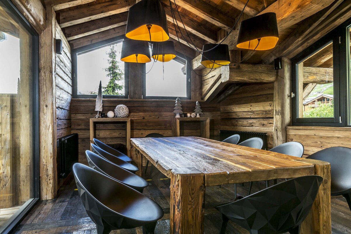 Cozy, rustic log cabin dining room with a large wooden table, sleek black chairs, and striking black & gold pendant lights, with mountain views.