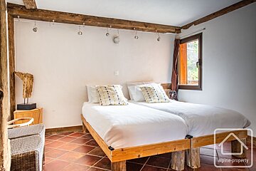 Rustic bedroom with two single beds, white bedding, exposed wooden beams, terracotta floor, and a window with a brown curtain.