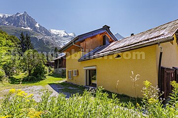 Bright yellow house with a rustic roof and wooden accents, backed by majestic snow-capped mountains and a verdant landscape under a clear sky.