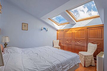 A light blue bedroom with a white bed, wooden wardrobe, and multiple skylights revealing a bright, cloudy sky. Two chairs sit by the wardrobe.