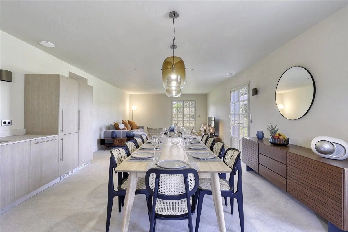 An open-concept dining room with a long wooden table, modern chairs, beige cabinets, and a living area in the background.