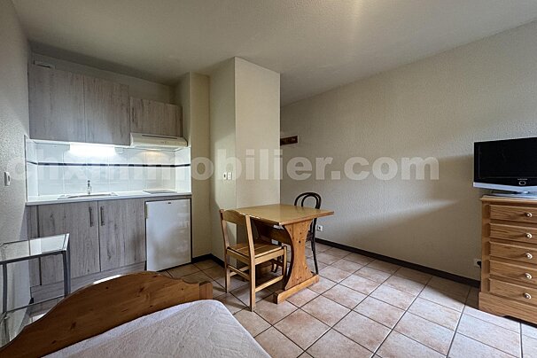 A compact studio apartment with a kitchenette, wooden dining table and chairs, a bed, and a TV on a dresser, all on a tiled floor.