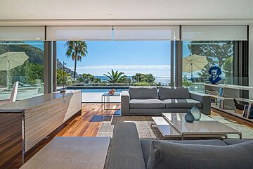 Bright modern living room with glass walls overlooking a pool, palm trees, and the sparkling sea. Features elegant gray sofas and wooden floors.