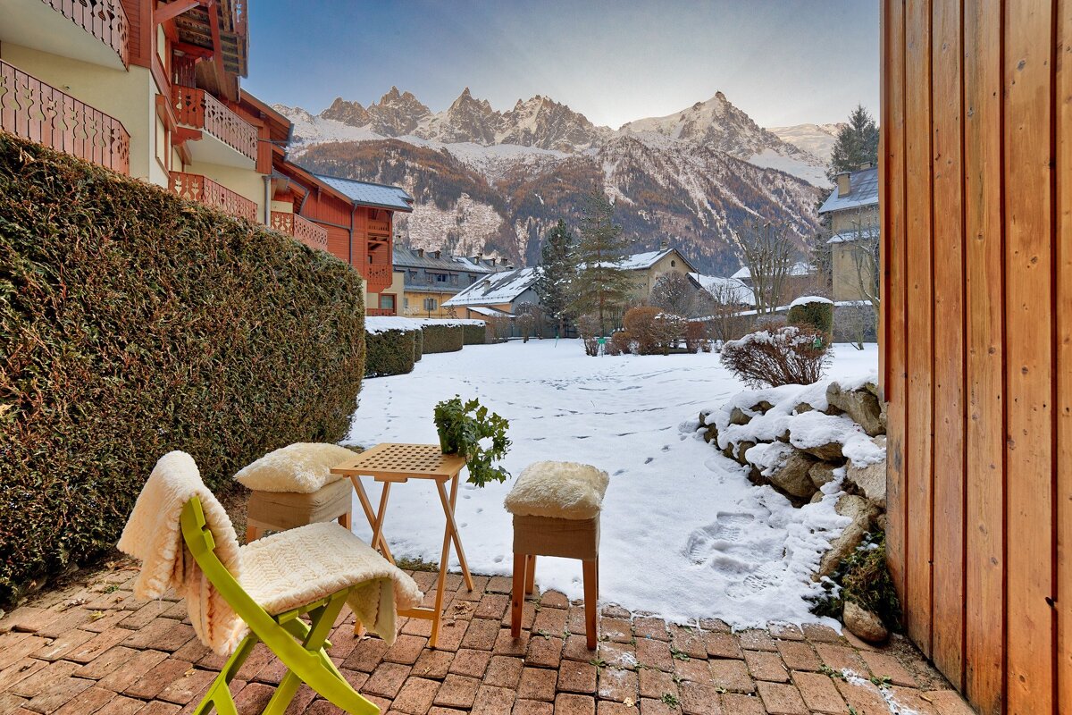 A cozy, brick patio with chairs and a table faces a snowy yard and village, all framed by towering, snow-capped mountains.
