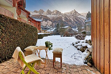 A cozy, brick patio with chairs and a table faces a snowy yard and village, all framed by towering, snow-capped mountains.