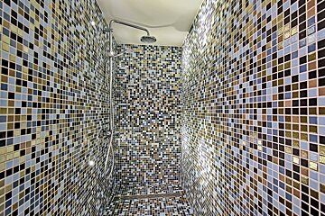 A modern shower stall lined with multi-colored blue, gold, and black mosaic tiles, complete with chrome fixtures.