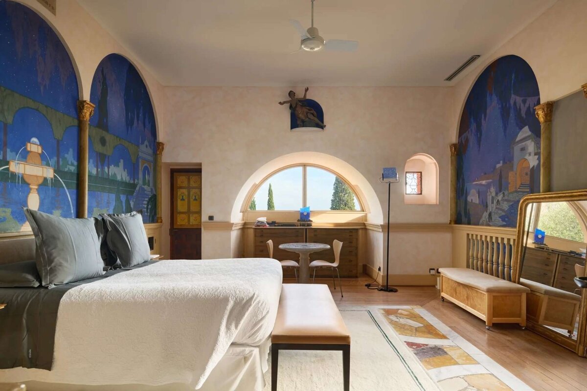 A lavish bedroom with dramatic blue arched murals, an arched window revealing a scenic view, and classic decor with a large bed.
