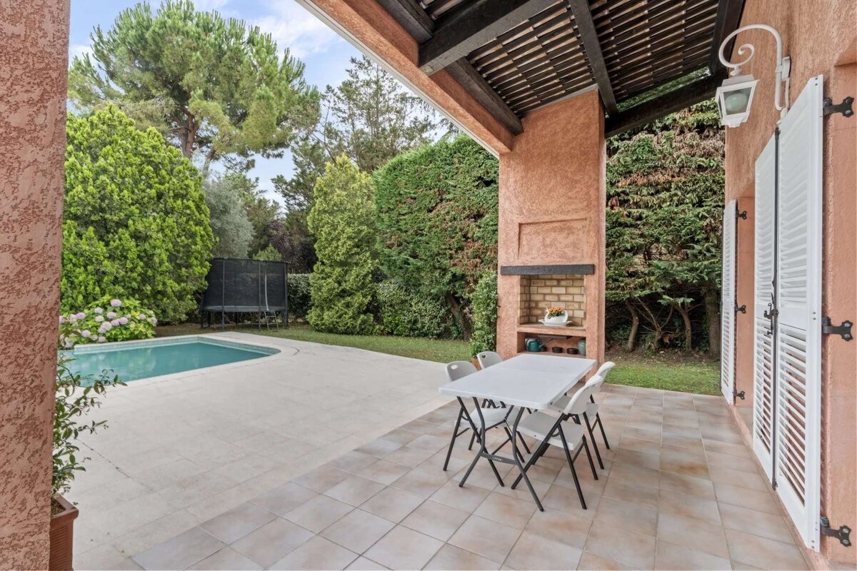 A spacious outdoor patio with a swimming pool, lush garden, dining table, and built-in BBQ next to a house with white shutters.