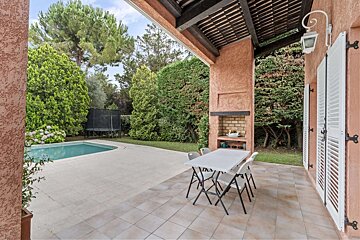 A spacious outdoor patio with a swimming pool, lush garden, dining table, and built-in BBQ next to a house with white shutters.