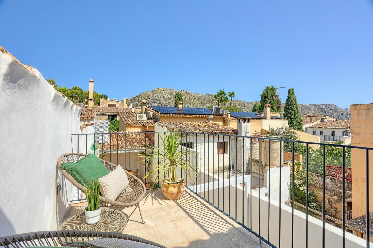 A sunny rooftop terrace with modern seating, plants, and a view over old town buildings, distant mountains, and a clear blue sky.