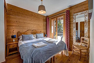 A cozy, rustic bedroom with wood paneled walls, a double bed with blue bedding, and glass doors leading to a balcony view.