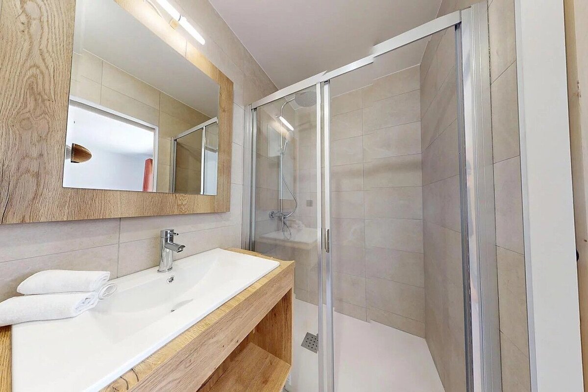 A modern bathroom features a wooden-framed mirror and vanity with a white sink, alongside a clear glass-enclosed shower, all in a neutral palette.