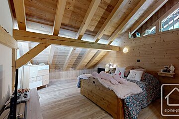 Cozy attic bedroom features extensive wood paneling, exposed beams, and a sloped ceiling. A bed with pink accents and a dresser create a warm, rustic vibe.