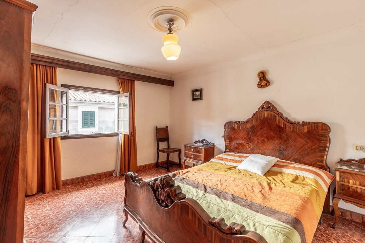 A vintage bedroom featuring an ornate wooden bed, striped bedding, an open window with orange curtains, and terracotta tiled floor.