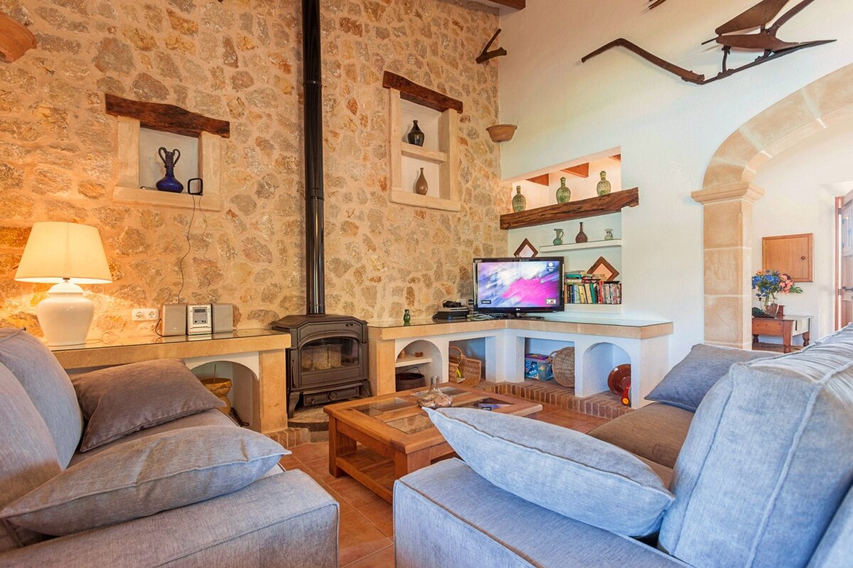 A cozy, rustic living room features textured stone walls, a black wood-burning stove, comfortable gray sofas, and a TV set up in a corner.