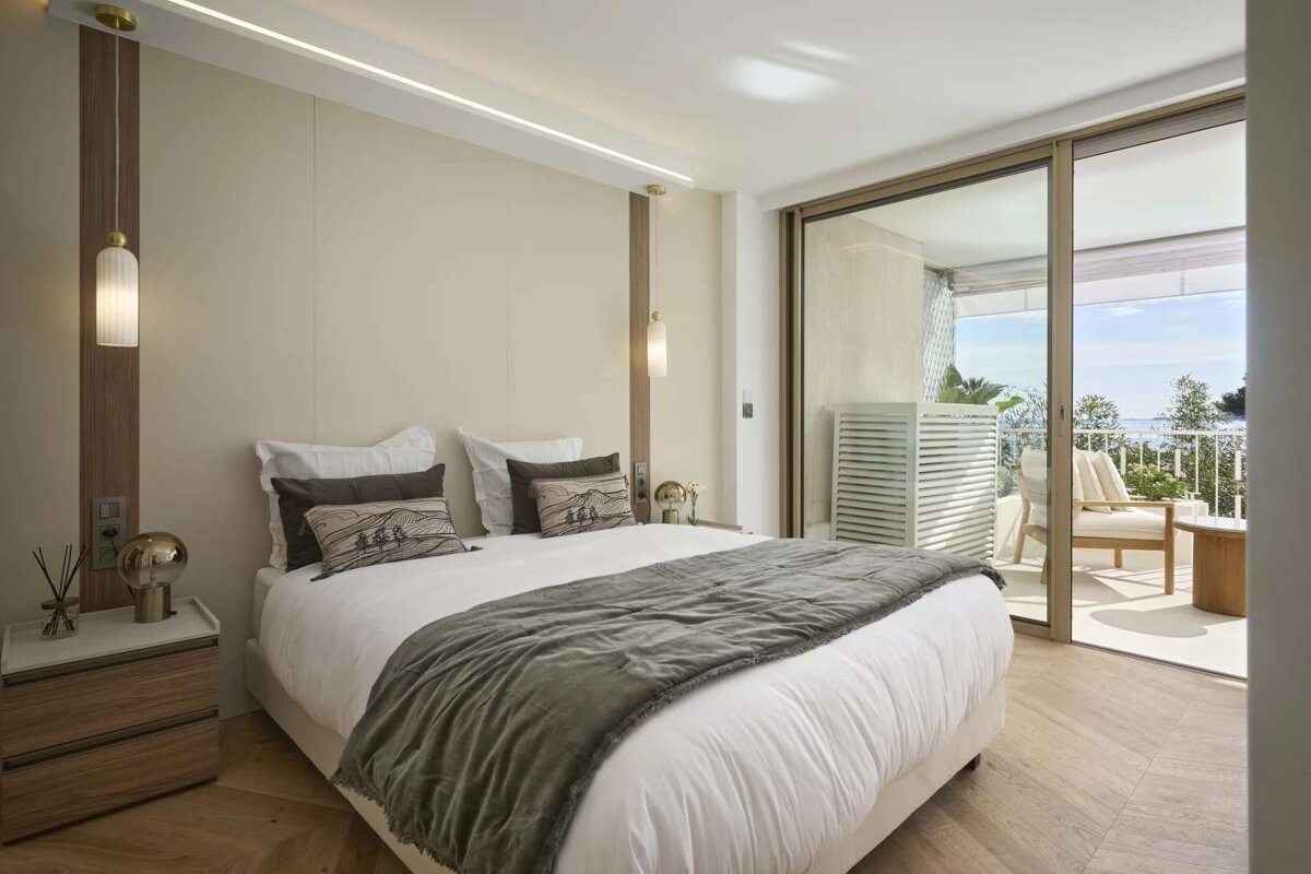 A chic bedroom in neutral tones with a king bed, wood accents, and sliding glass doors leading to a balcony with a beautiful sea view.