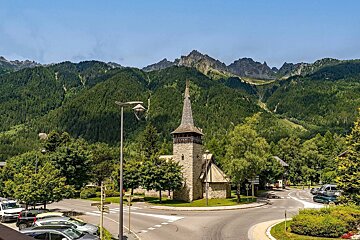 A stone church with a spire stands by a road and cars, surrounded by lush green trees and towering mountains under a clear blue sky.