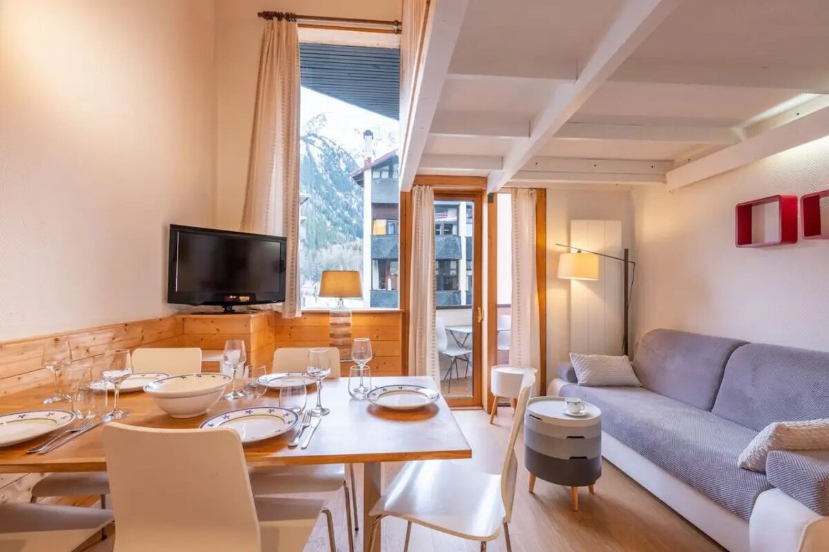 A cozy, modern apartment interior featuring a set dining table, grey sofa, and a window with mountain views.