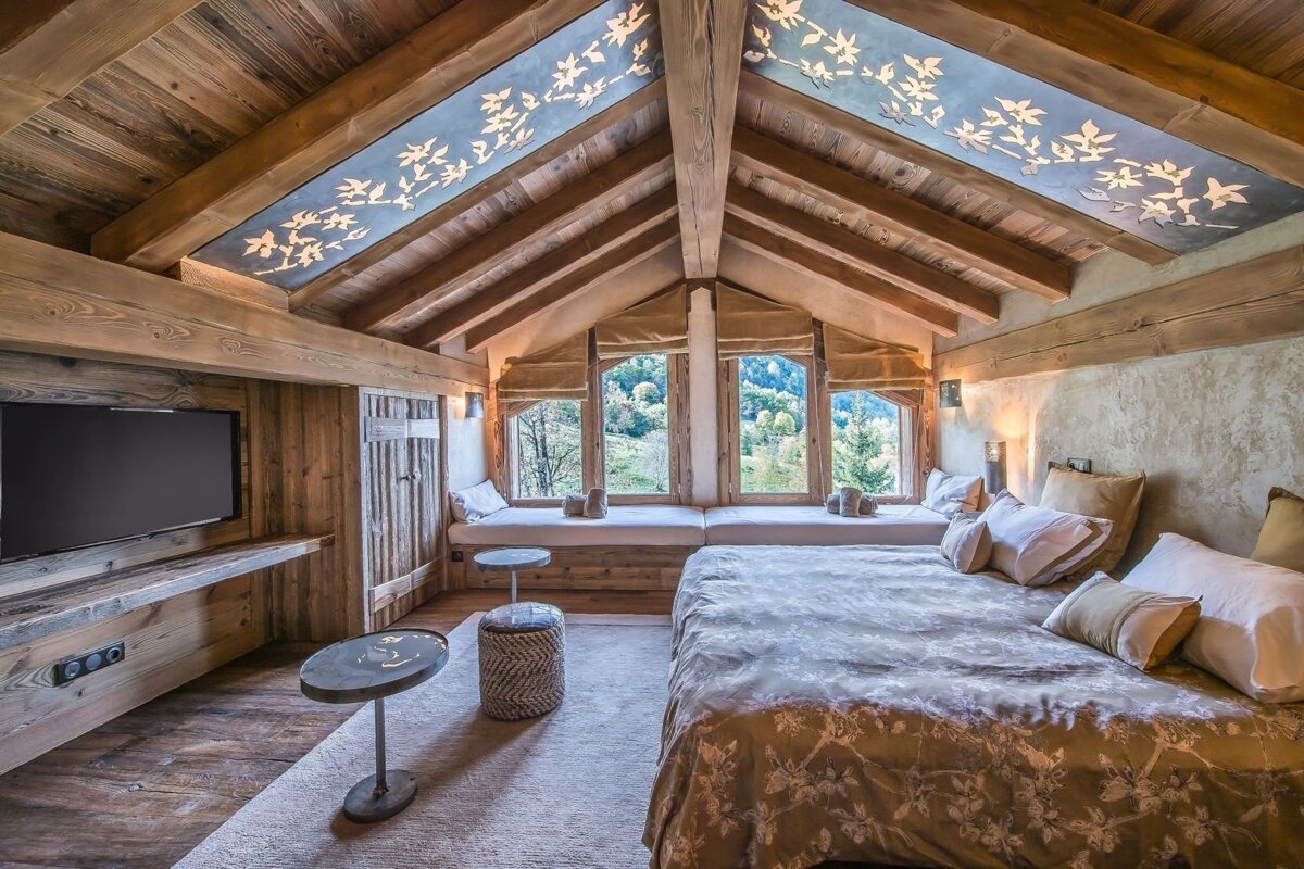 A rustic attic bedroom featuring exposed wood beams, an illuminated ceiling with leaf patterns, a cozy bed, and large windows offering scenic mountain views.