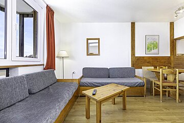 A bright living/dining room with grey sofas, wooden coffee table, and a round dining set. Features white walls, warm wood accents, and a large window.