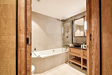Modern bathroom with a grey tiled bathtub, a wooden vanity with a vessel sink, and a mirror, framed by a rustic wooden doorway.