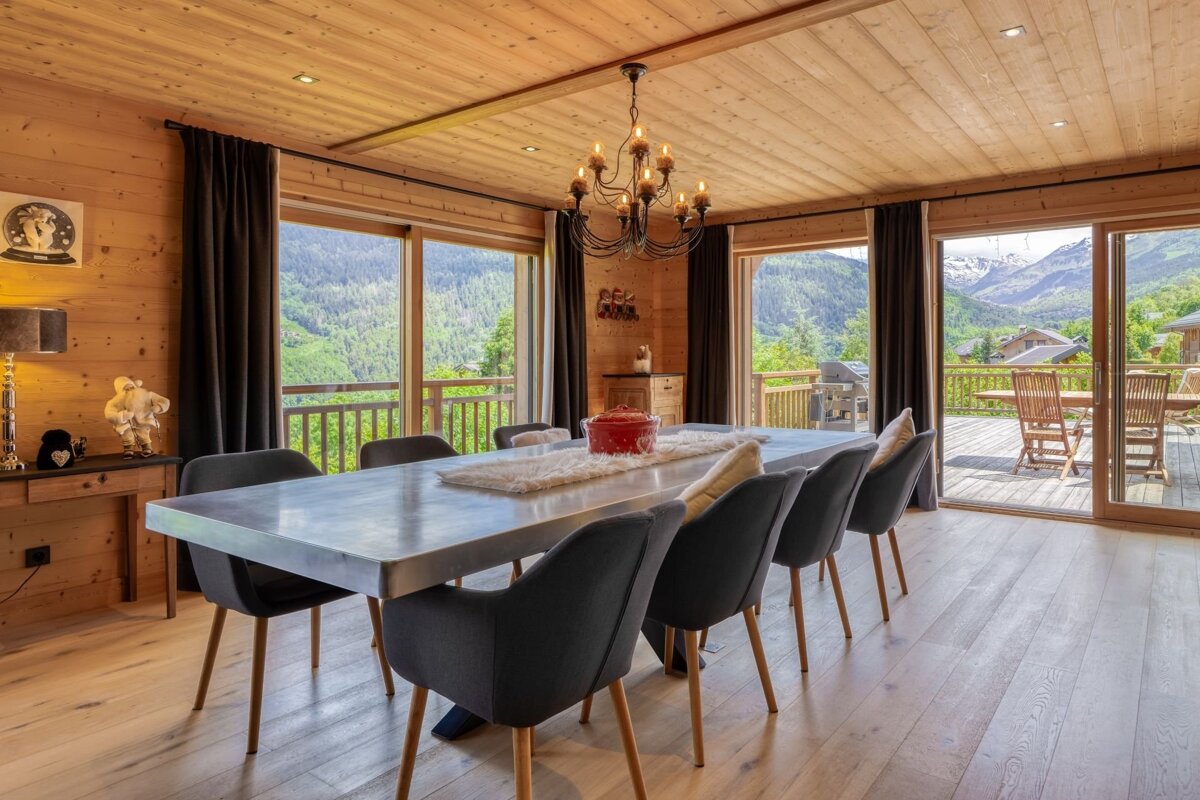 A rustic wooden dining room with a long table, chandelier, and expansive windows offering stunning mountain and forest views.