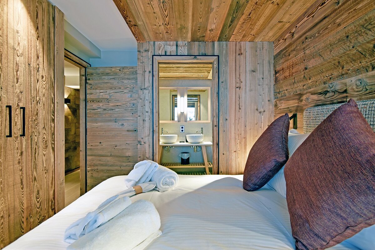 Chalet-style bedroom with a bed, white towels, and brown pillows. A modern bathroom with double sinks is visible through a doorway.