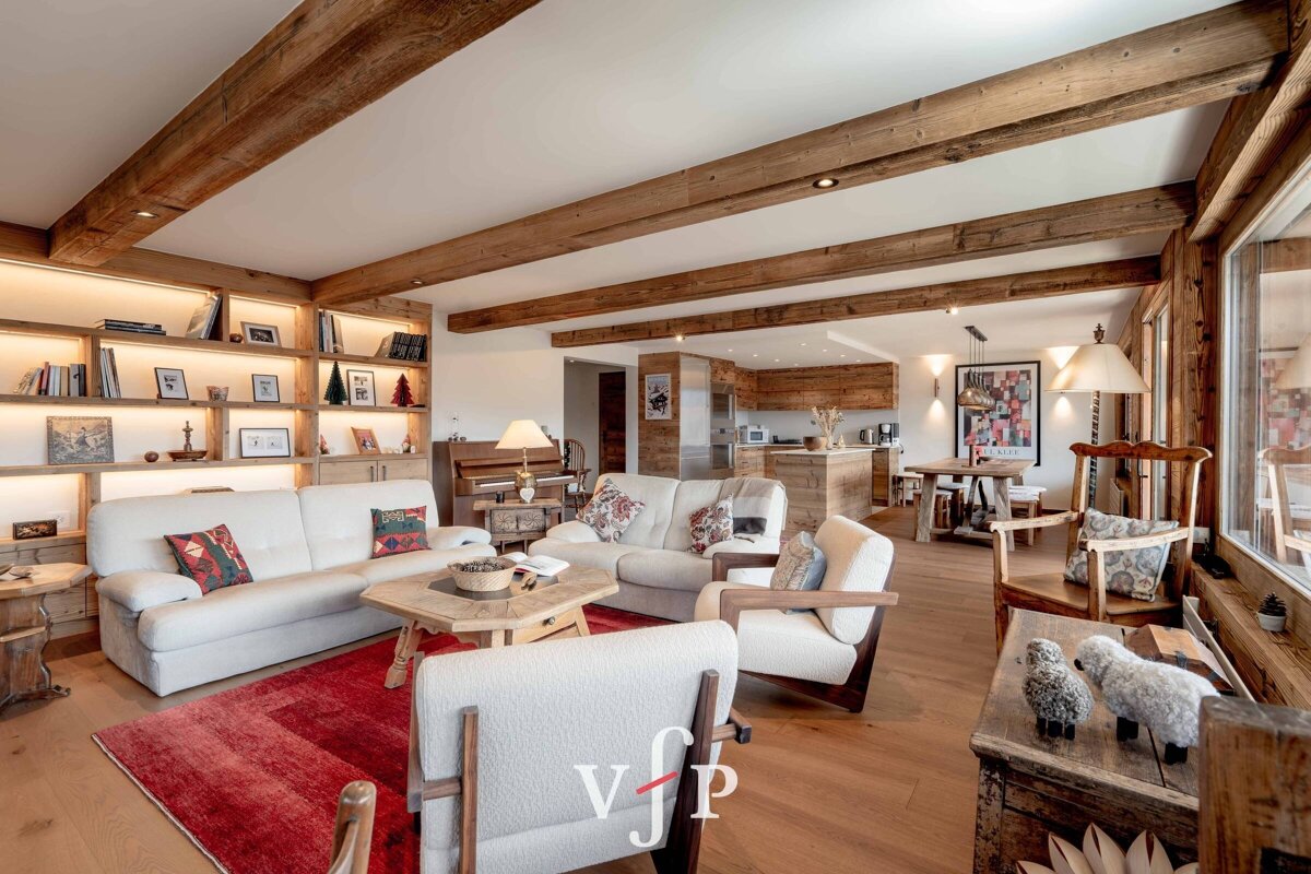 Cozy alpine chalet living room with exposed wood ceiling beams, white sofas on a red rug, and a kitchen visible in the background.