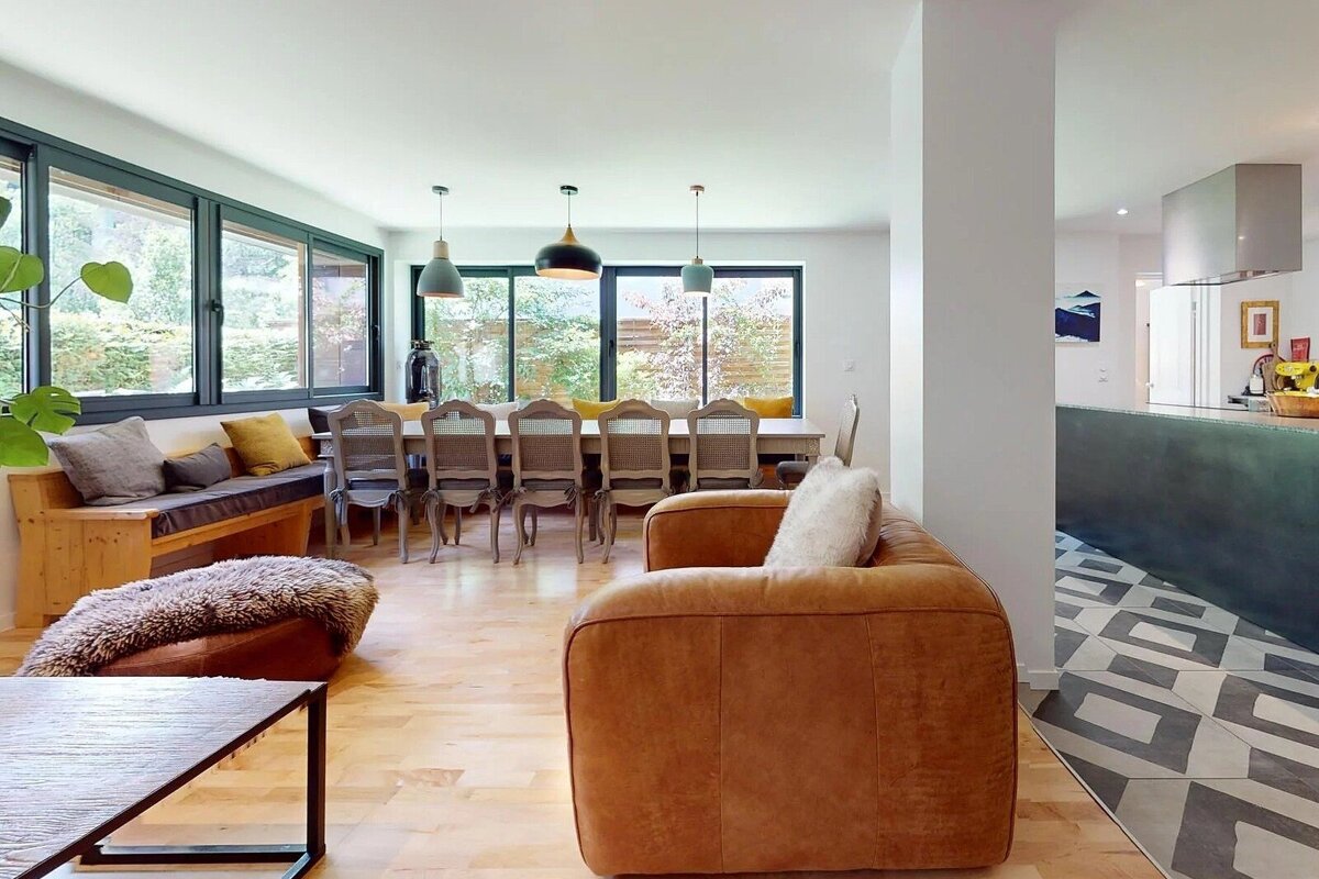 Bright, open-plan living & dining room with a tan leather sofa, long dining table, window bench, wooden floors, and a modern kitchen.