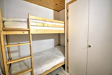 A compact room features wooden bunk beds with white bedding, a wood-paneled ceiling, white walls, and a white closet/door on the right.