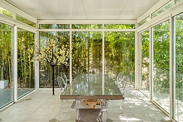 A modern, light-filled dining room with glass walls offering panoramic views of a lush green bamboo forest. Features a glass table and clear chairs.
