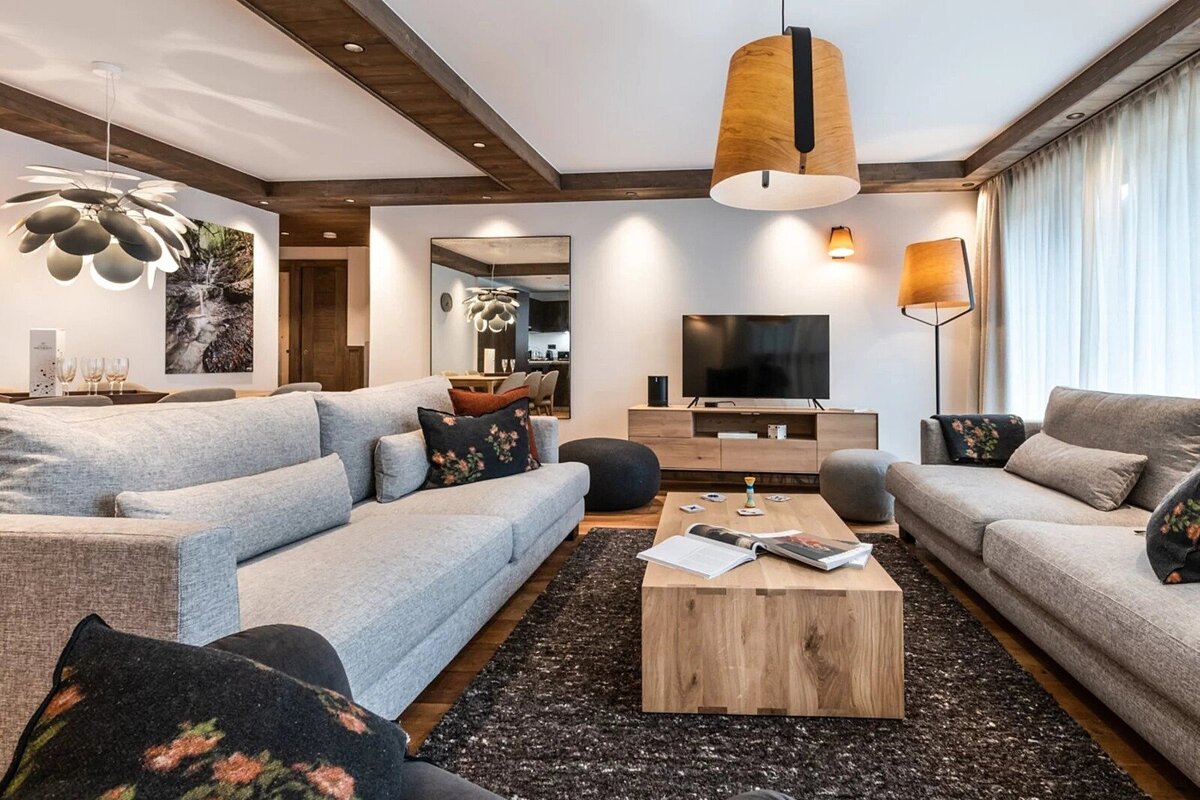 A stylish, modern living room with large grey sofas, a wooden coffee table, and distinctive lighting. Exposed wooden beams add warmth and character.