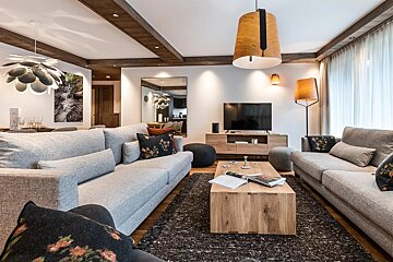 A stylish, modern living room with large grey sofas, a wooden coffee table, and distinctive lighting. Exposed wooden beams add warmth and character.