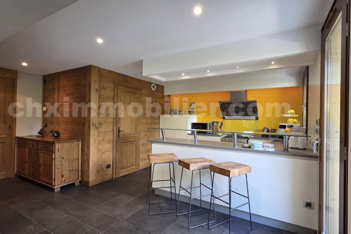 A modern kitchen with a breakfast bar, three stools, and bright orange accents. Rustic wooden walls complement sleek white counters and dark tiled floor.