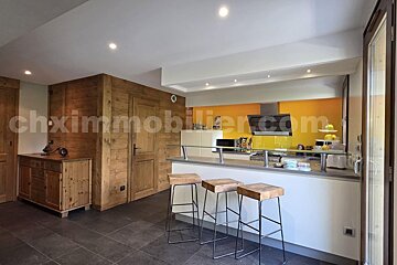 A modern kitchen with a breakfast bar, three stools, and bright orange accents. Rustic wooden walls complement sleek white counters and dark tiled floor.