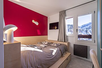 Modern bedroom with a red accent wall, wooden platform bed, and a TV. A window offers a beautiful view of snow-capped mountains.