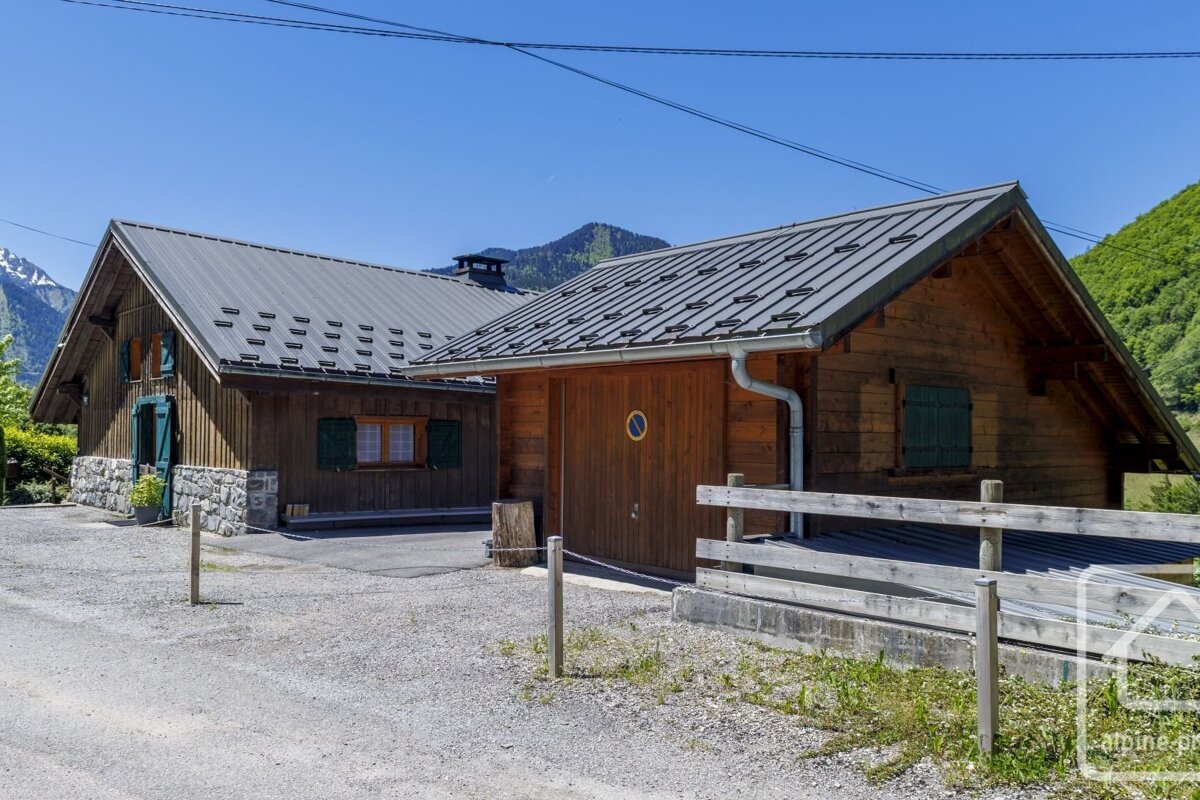 Two rustic wooden chalets with metal roofs under a clear blue sky, set against a backdrop of green mountains and snowy peaks.