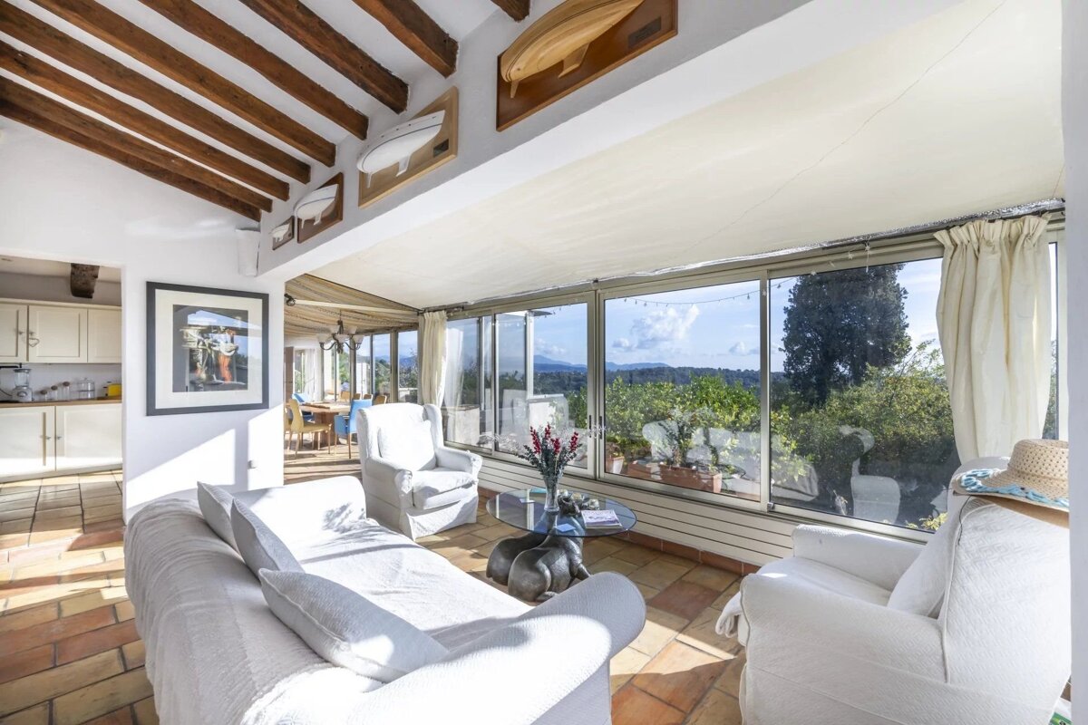 A bright, sunlit living room with white sofas, wooden ceiling beams, and large windows overlooking a scenic green landscape.