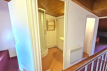 A small bathroom with a corner shower, wooden ceiling, and shelves, viewed from a hallway with a white textured wall.