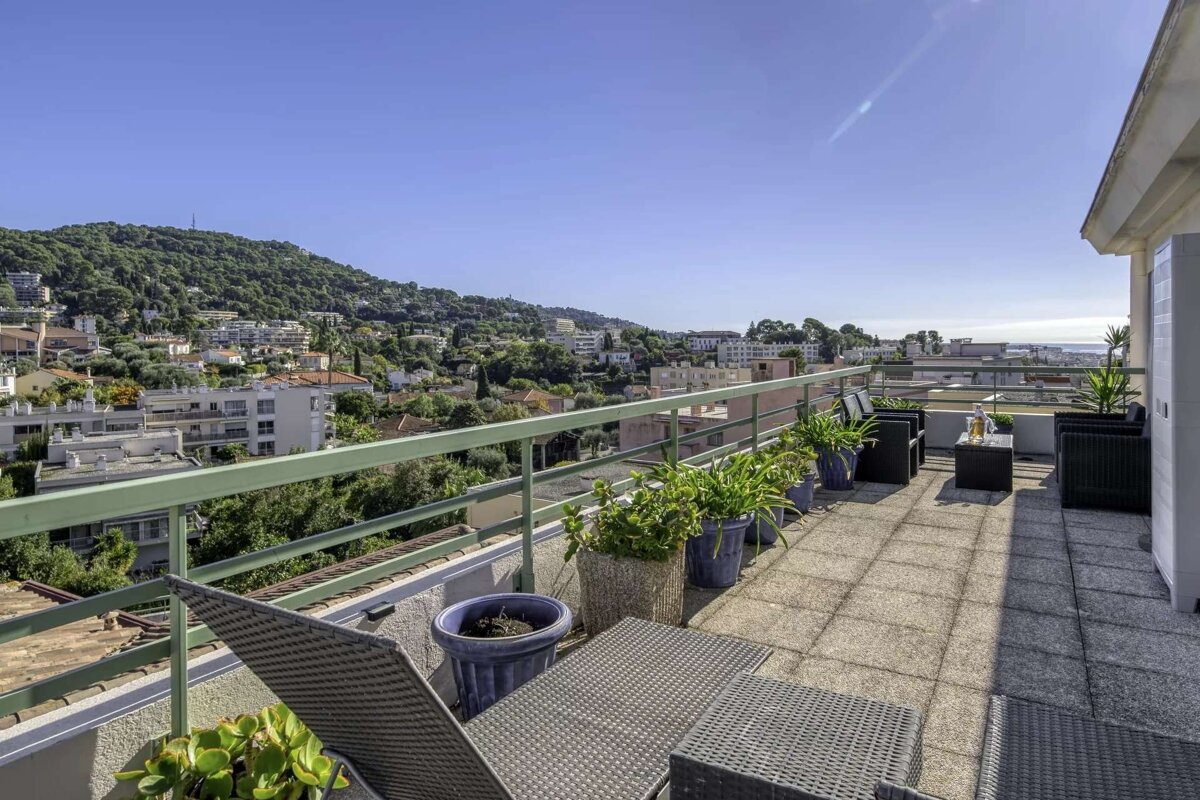 Sunny rooftop terrace with lounge furniture and plants, offering panoramic views of a hillside town, lush greenery, and the distant sea.