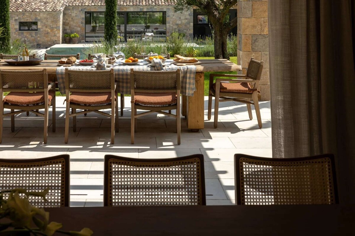 A rustic outdoor dining table, set with food, sits on a sunlit patio with woven chairs. A stone house and garden provide a serene backdrop.
