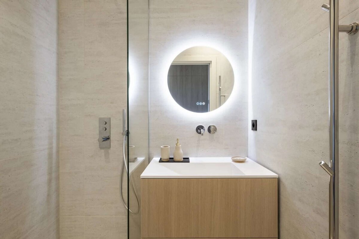 A modern, neutral-toned bathroom with light tiles features a round backlit mirror above a wood vanity. A glass panel separates the shower area.