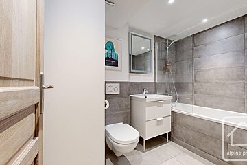 A modern bathroom with grey tiles, a white vanity, a toilet, and a bathtub with a glass screen.
