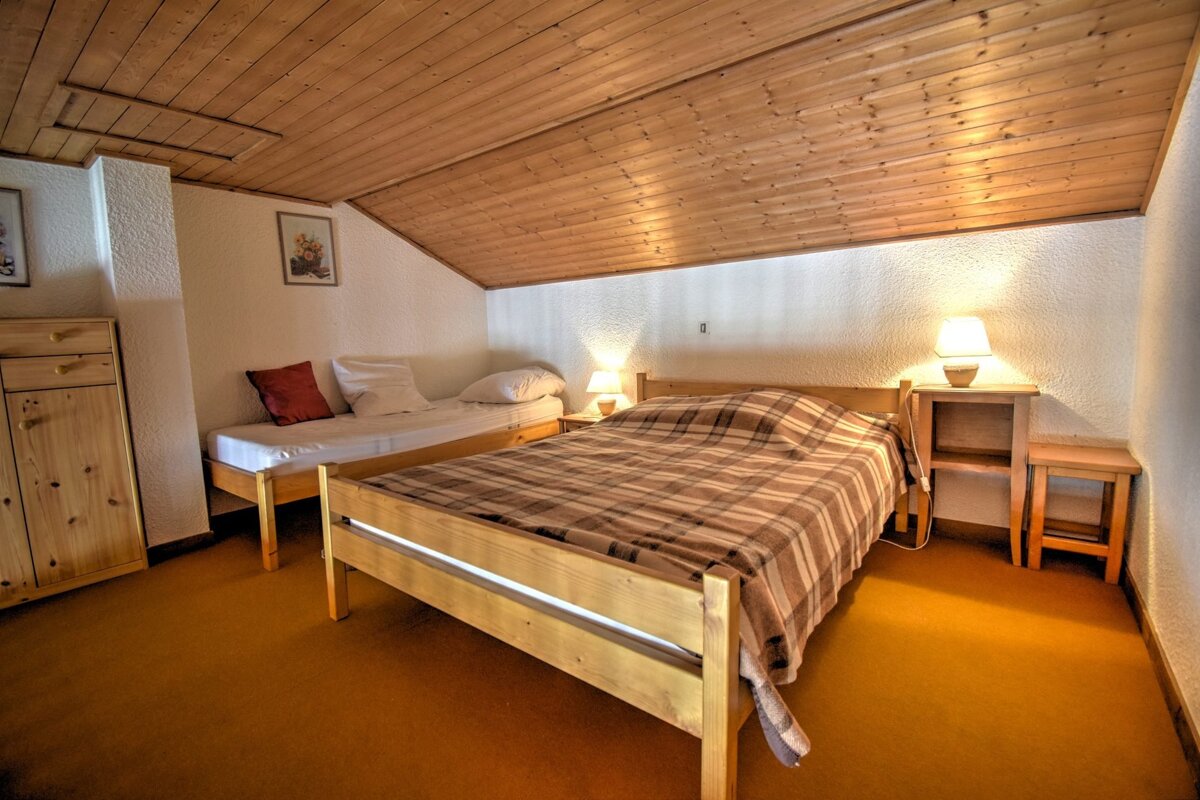 Cozy attic bedroom with a sloped wooden ceiling, featuring a double bed with a plaid blanket, a single bed, and light wood furniture on an orange carpet.