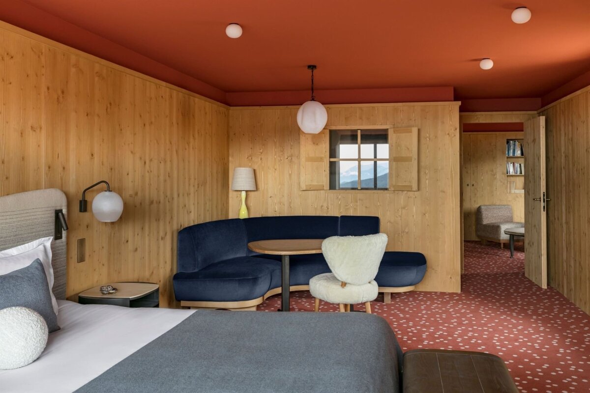 A cozy, wood-paneled room with a red ceiling, patterned carpet, and a blue curved sofa. A bed is in the foreground and a window shows mountain views.