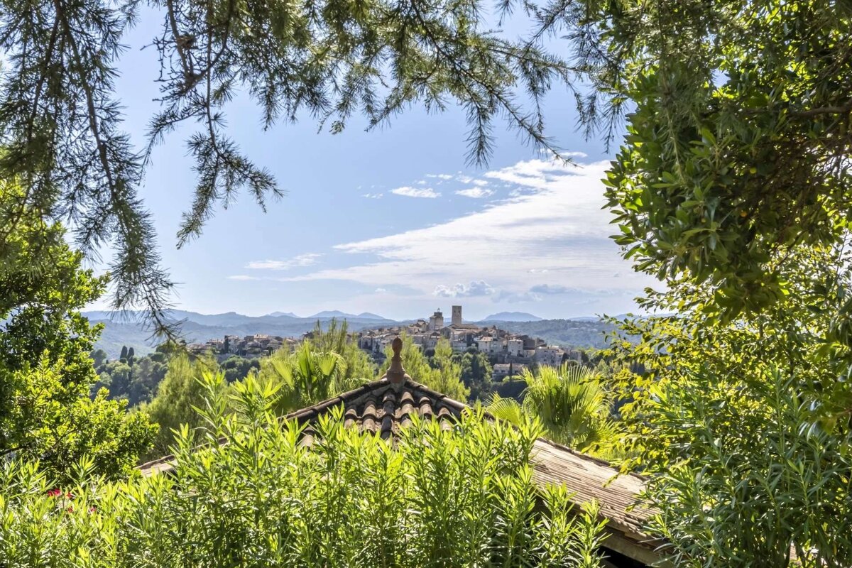 A vibrant green landscape frames a distant medieval hill town with prominent towers, under a bright, cloudy sky, with a tiled roof visible.